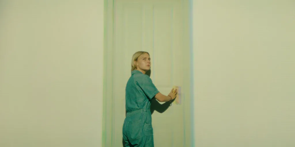 a blonde girl in overalls in a white room going through a door and looking over her shoulder