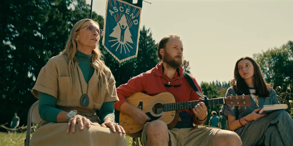 A blonde woman (Toni Collette) alongside a man playing guitar and another woman, all at a camp.