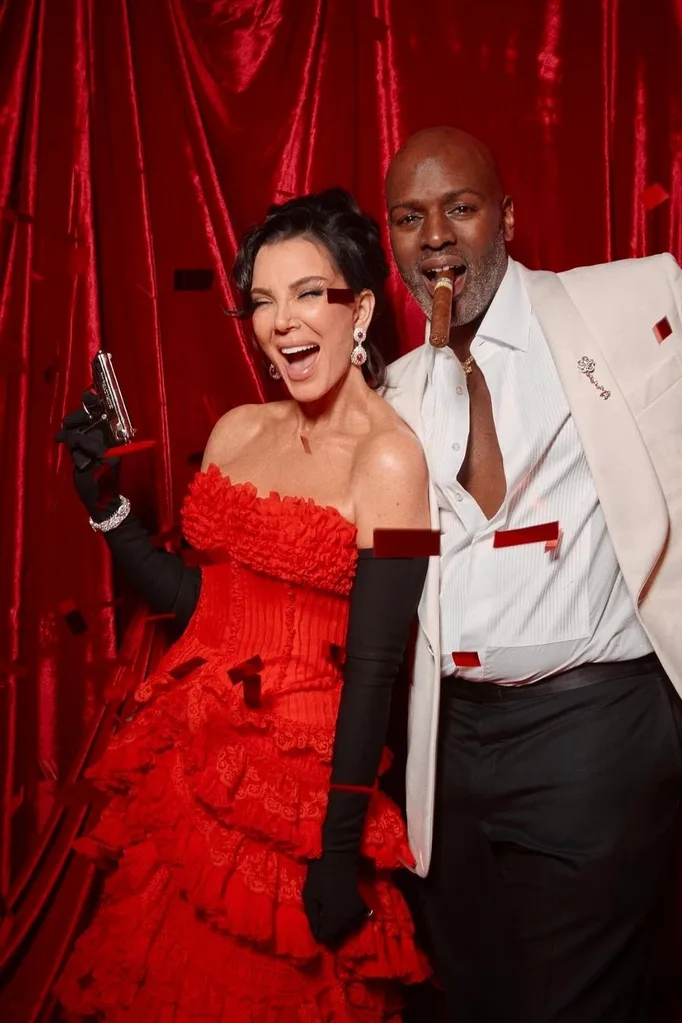 kris jenner in a red dress holding a prop gun alongside boyfriend Corey Gamble in a white tux and cigar in his mouth, both are smiling and attending kris' party.