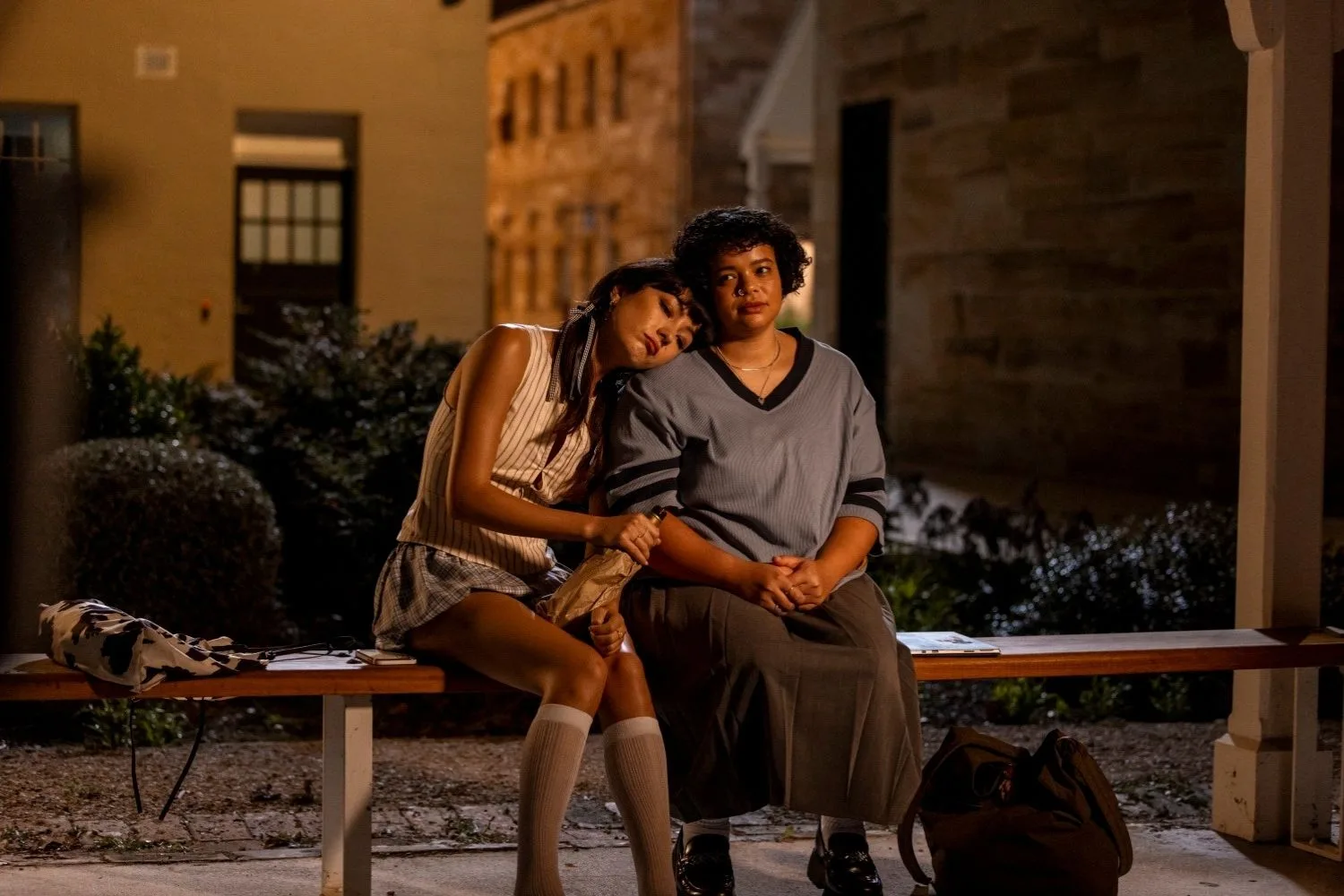 two girls at night sitting on a bench, the girl on the left is resting her head on the other's shoulder, a scene from he had it coming