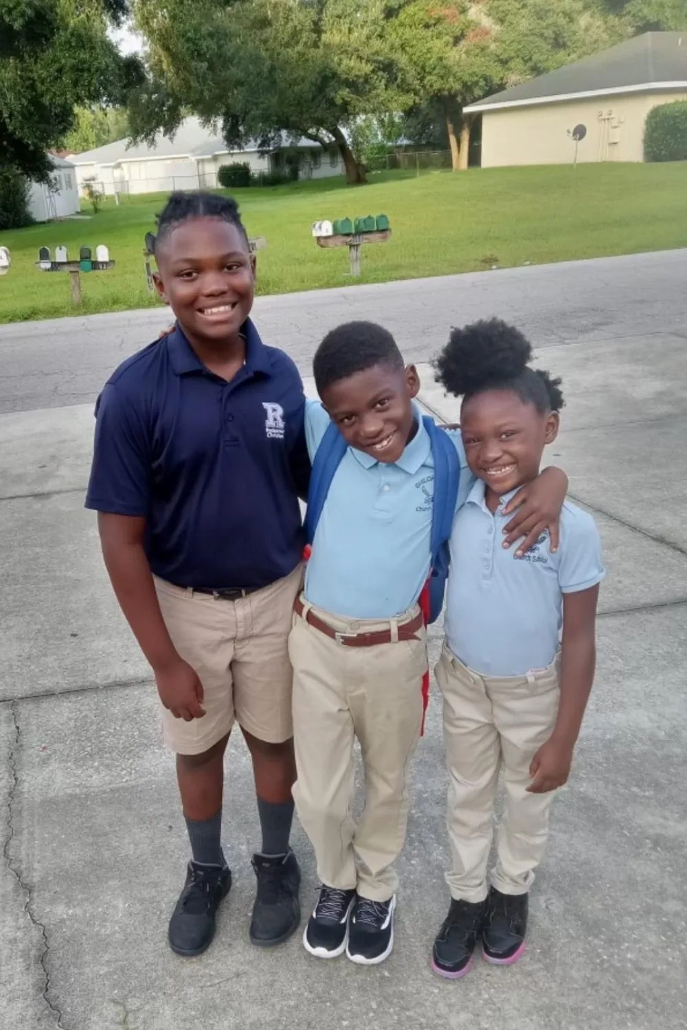 three young children posing together in a driveway with their school uniforms on, smiling