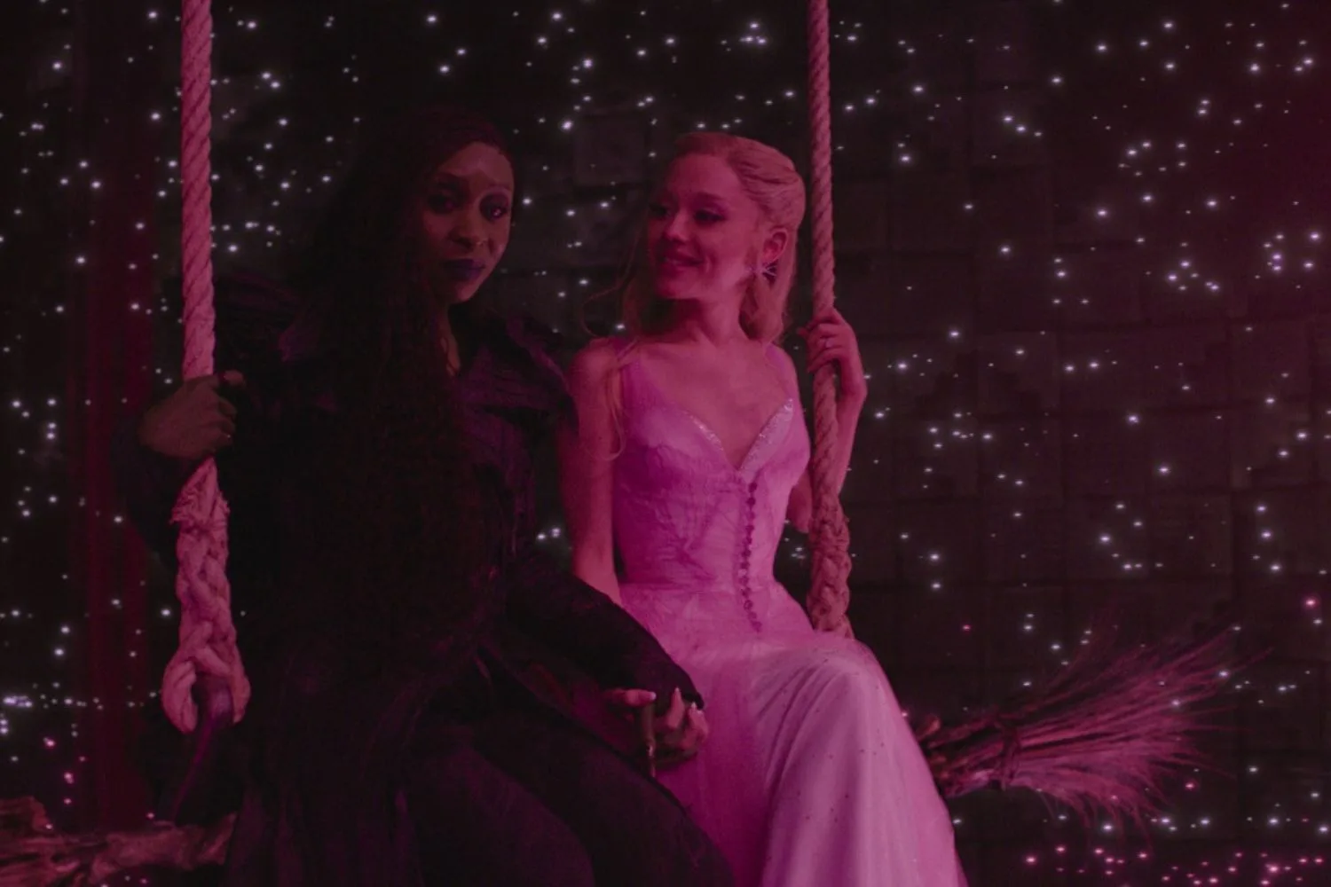 elphaba and glinda sitting on the broomstick in a darkly lit room with hot pink lighting