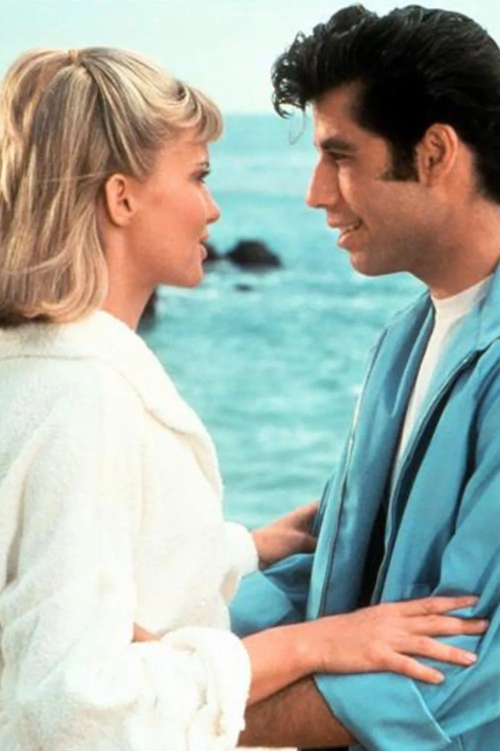 Woman with blonde hair in white shirt staring into a man with brown hair wearing a blue shirt at the beach portraying a summer romance