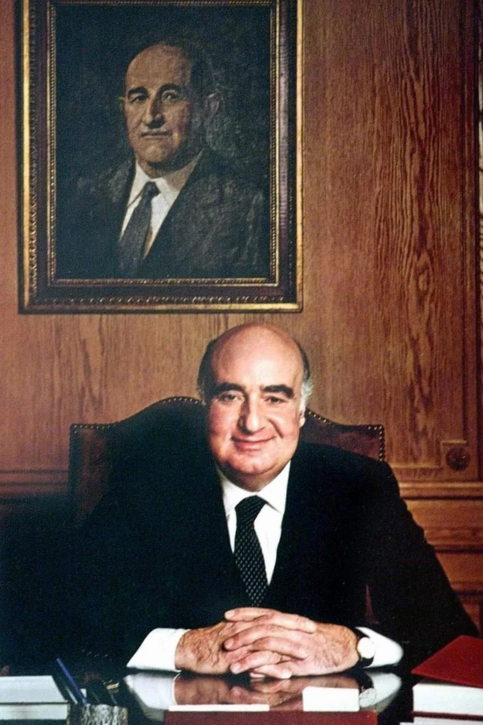 edmond safra, an older man sitting at a desk with a painting of himself behind him on the wall