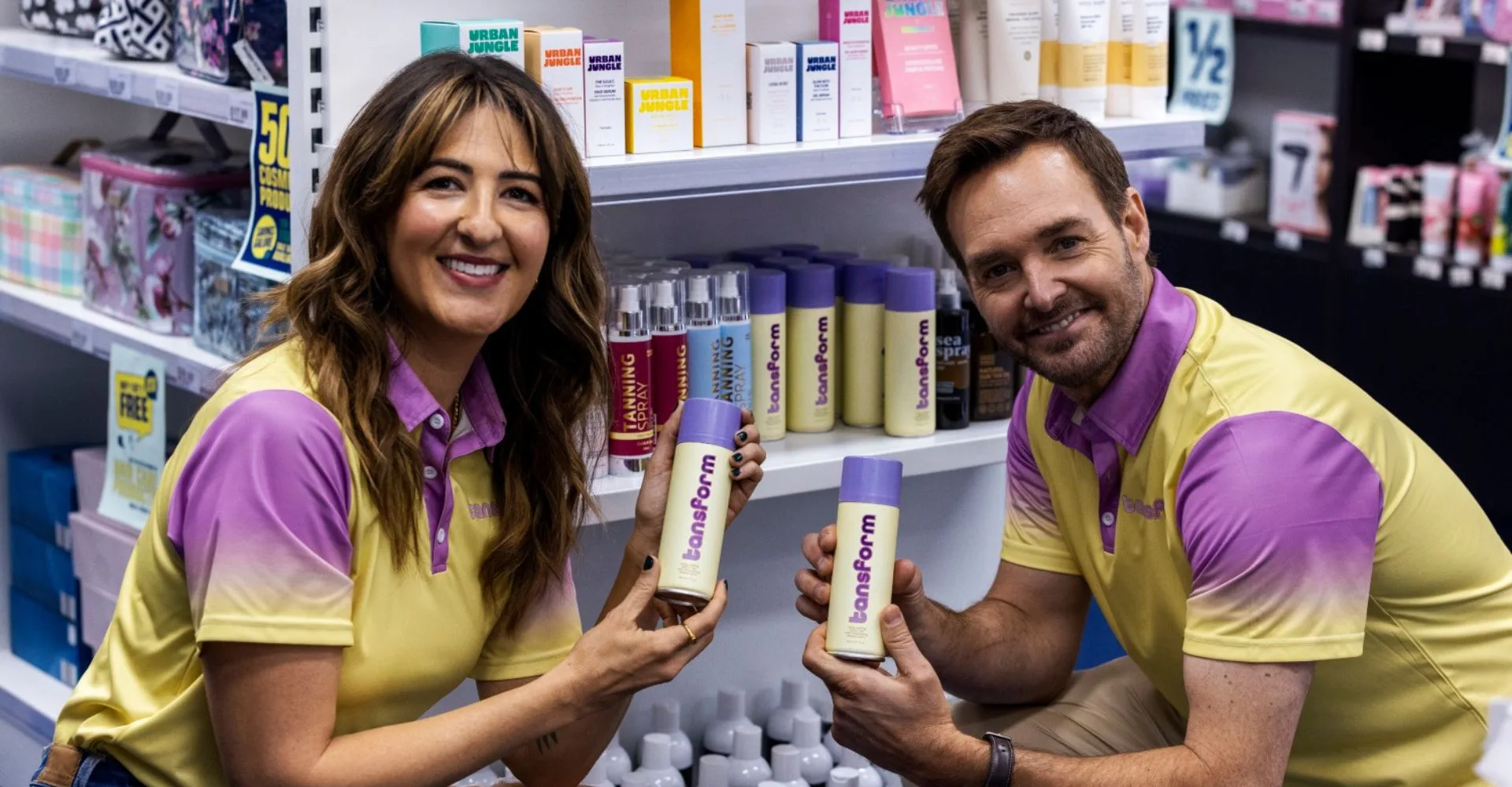 d'arcy carden and will forte posing in character with their characters' tan product in a chemist in yellow and purple uniforms