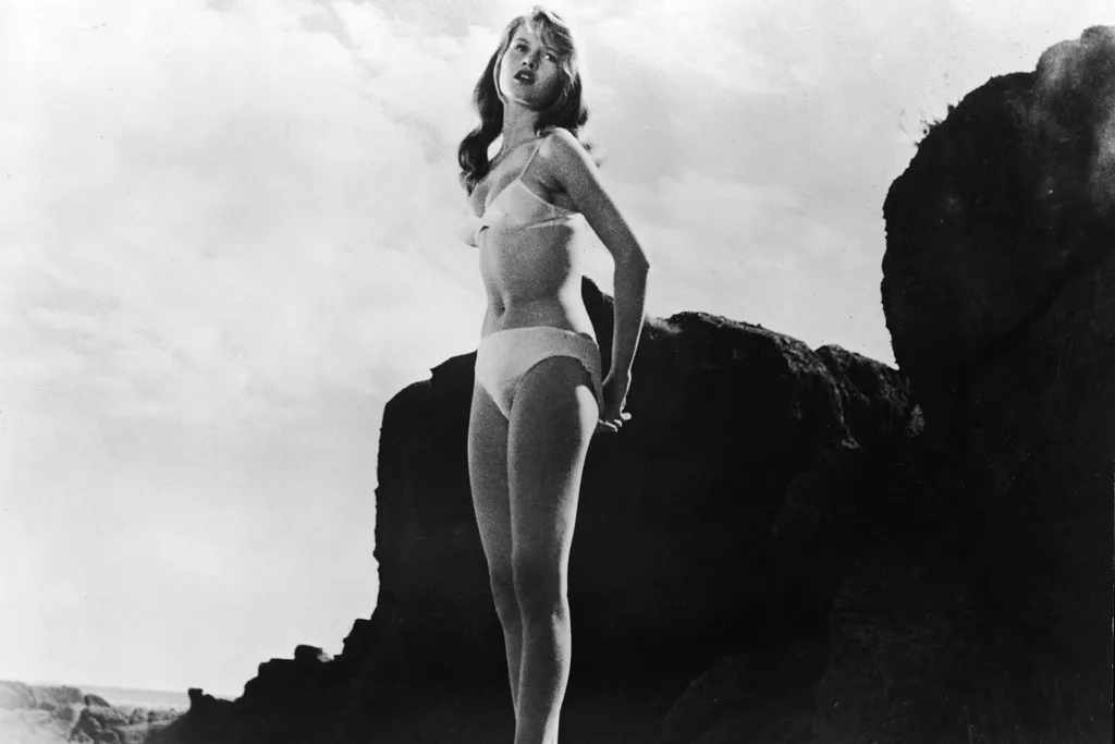 French-born actor Brigitte Bardot wears a white bikini and stands on a rocky beach in a still from the film, 'The Girl in the Bikini', directed by Willy Rozier, 1958.