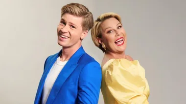 Robert Irwin wears a blue suit and Julia Morris wears a yellow dress. The pair smile back-to-back.