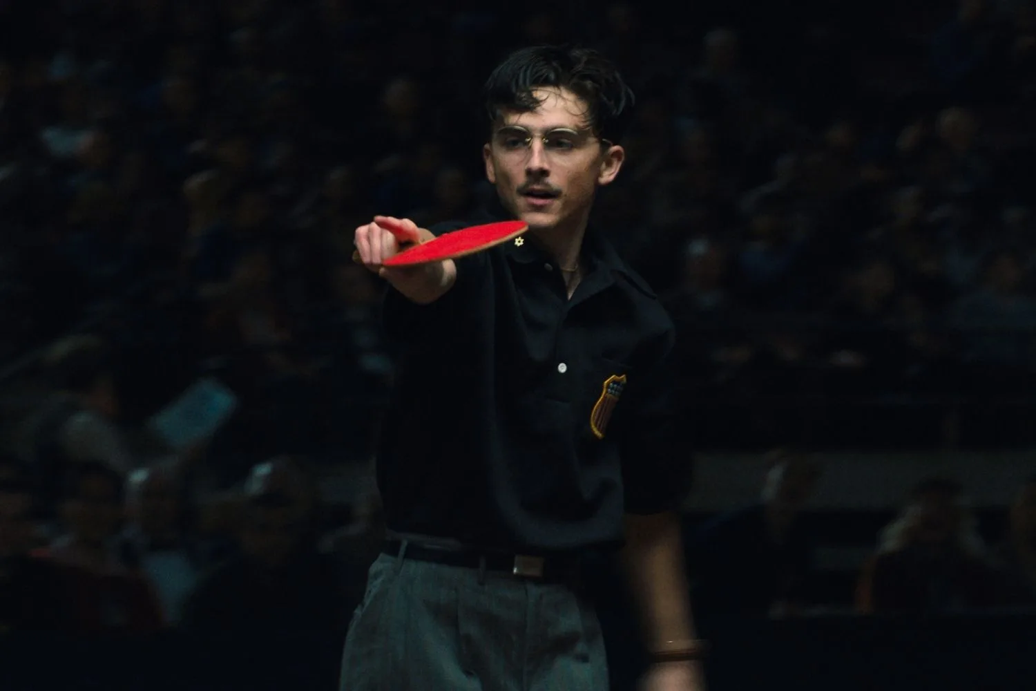 timothee chalamet in character playing table tennis with glasses on in a dark room