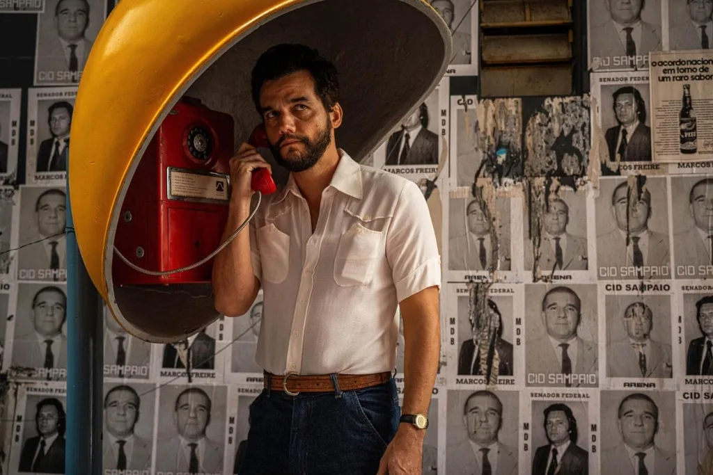 wagner moura in a button up shirt and jeans talking on the phone at a phone booth