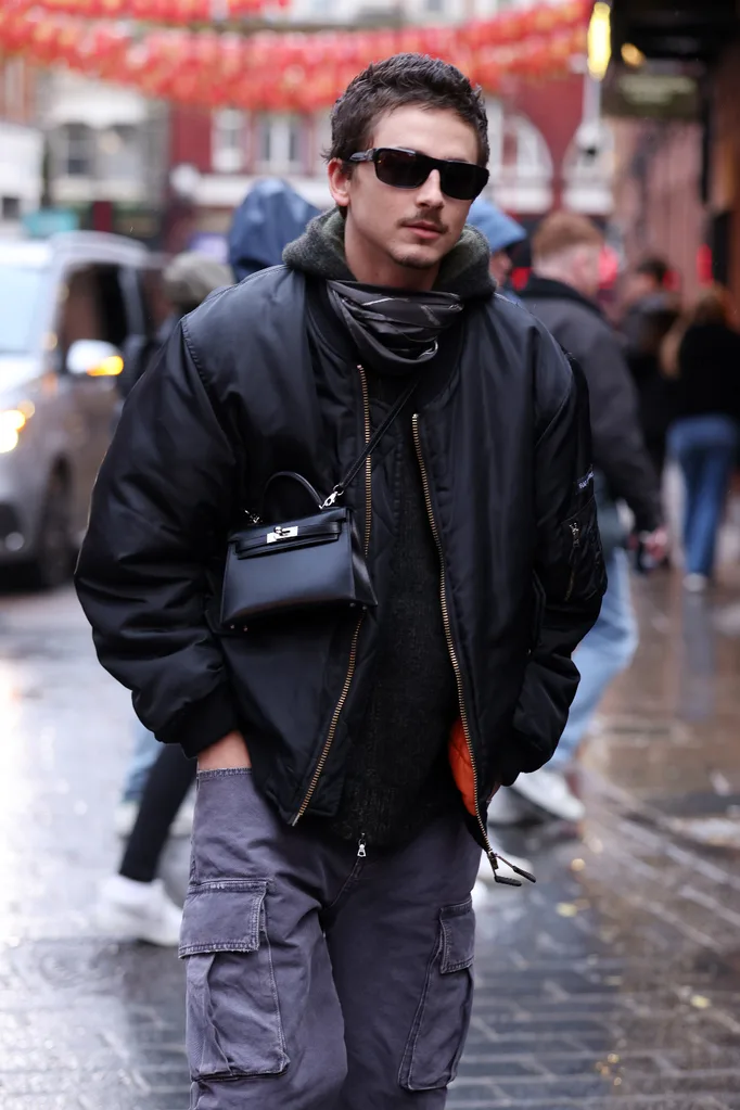 Timothee Chalamet wearing a black jacket and grey pants, accessorising with a small crossbody "man bag"