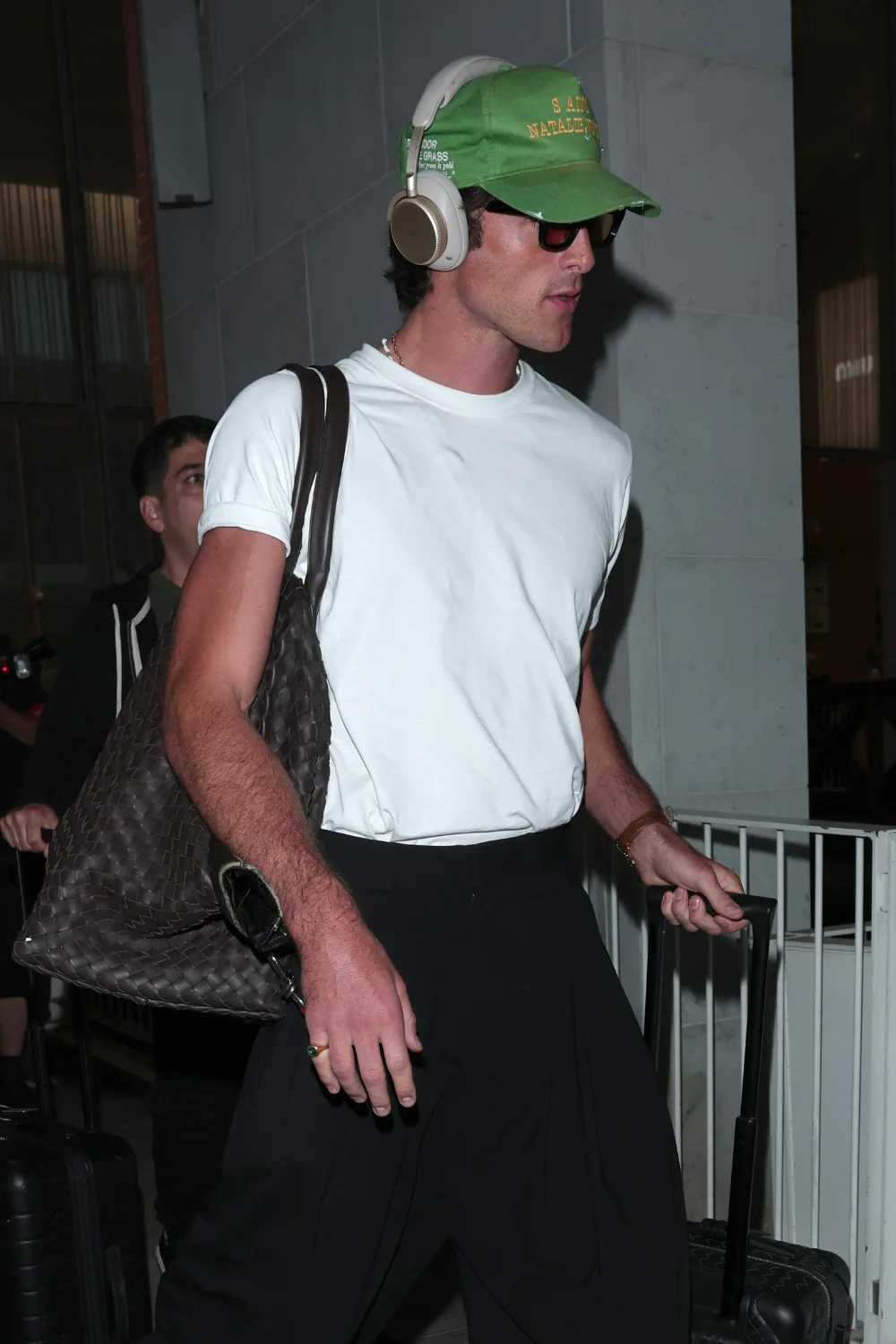 Jacob Elordi wearing a white t-shirt,black pants, a green cap and headphones carrying a woven-look "man bag"