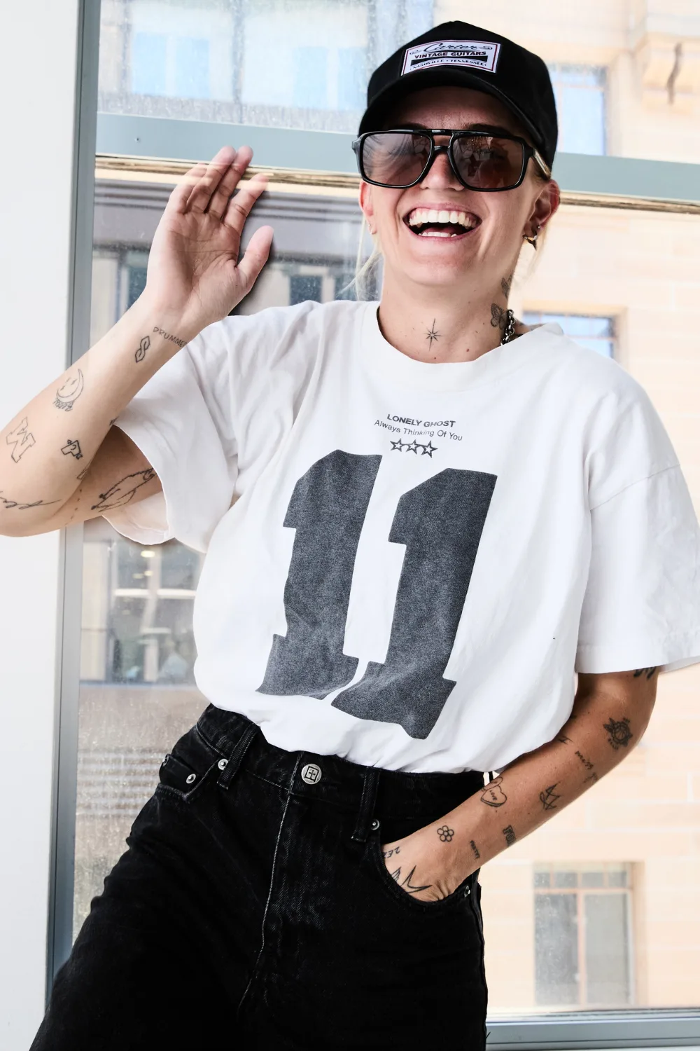 G Flip smiling wearing a cap, sunglasses, a white t-shirt with an 11 printed on it and black jeans.