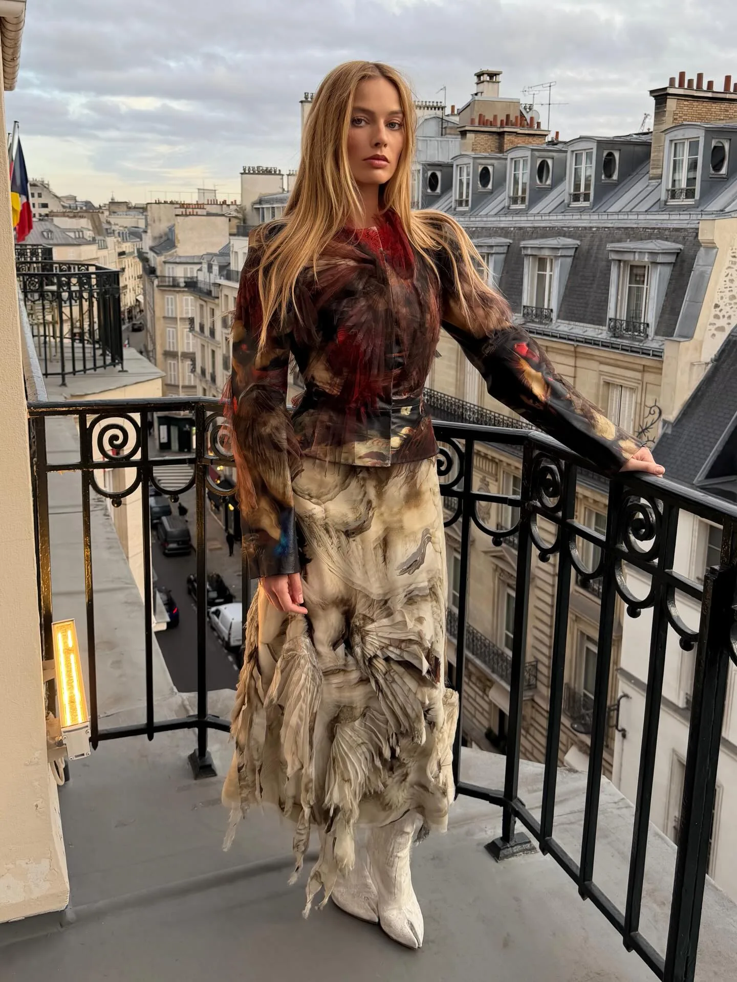 Margot Robbie wuthering heights style: Margot Robbie a brown jacket and cream skirt standing overlooking a balcony. 