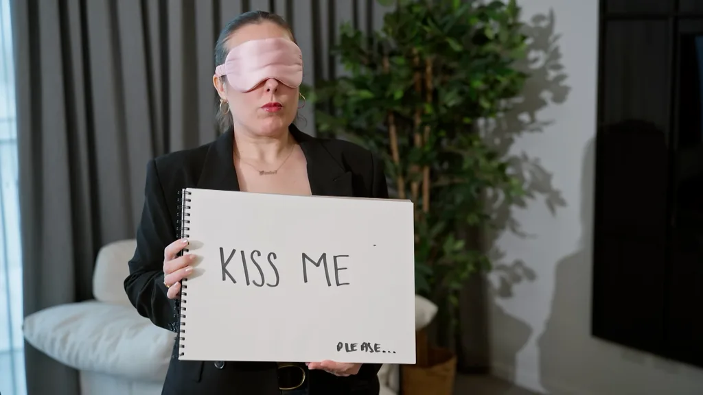 rachel standing waiting for steven while she wears a blindfold and hold a sign that says 'kiss me'