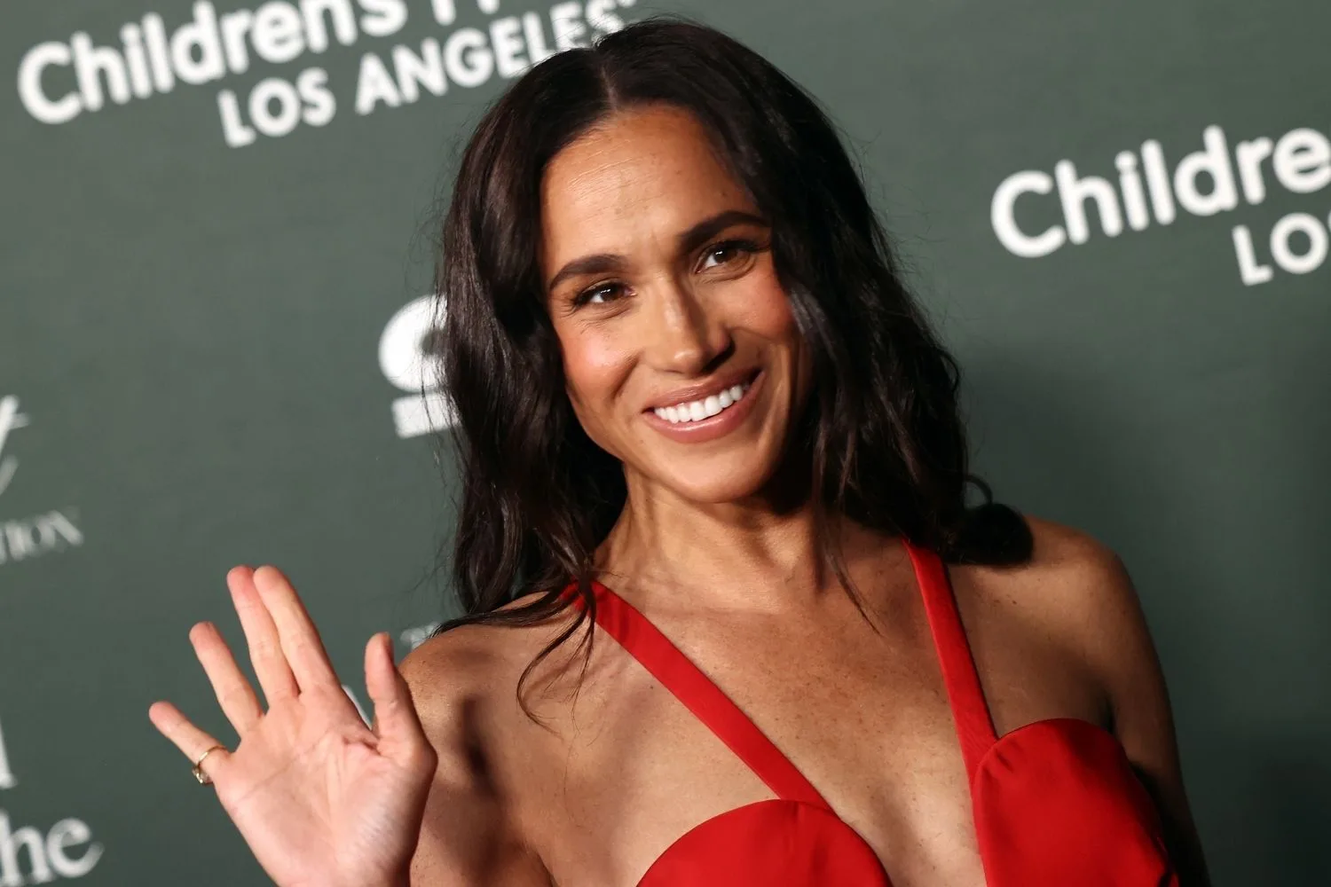 Meghan Markle smiling and waving wearing a red dress. 
