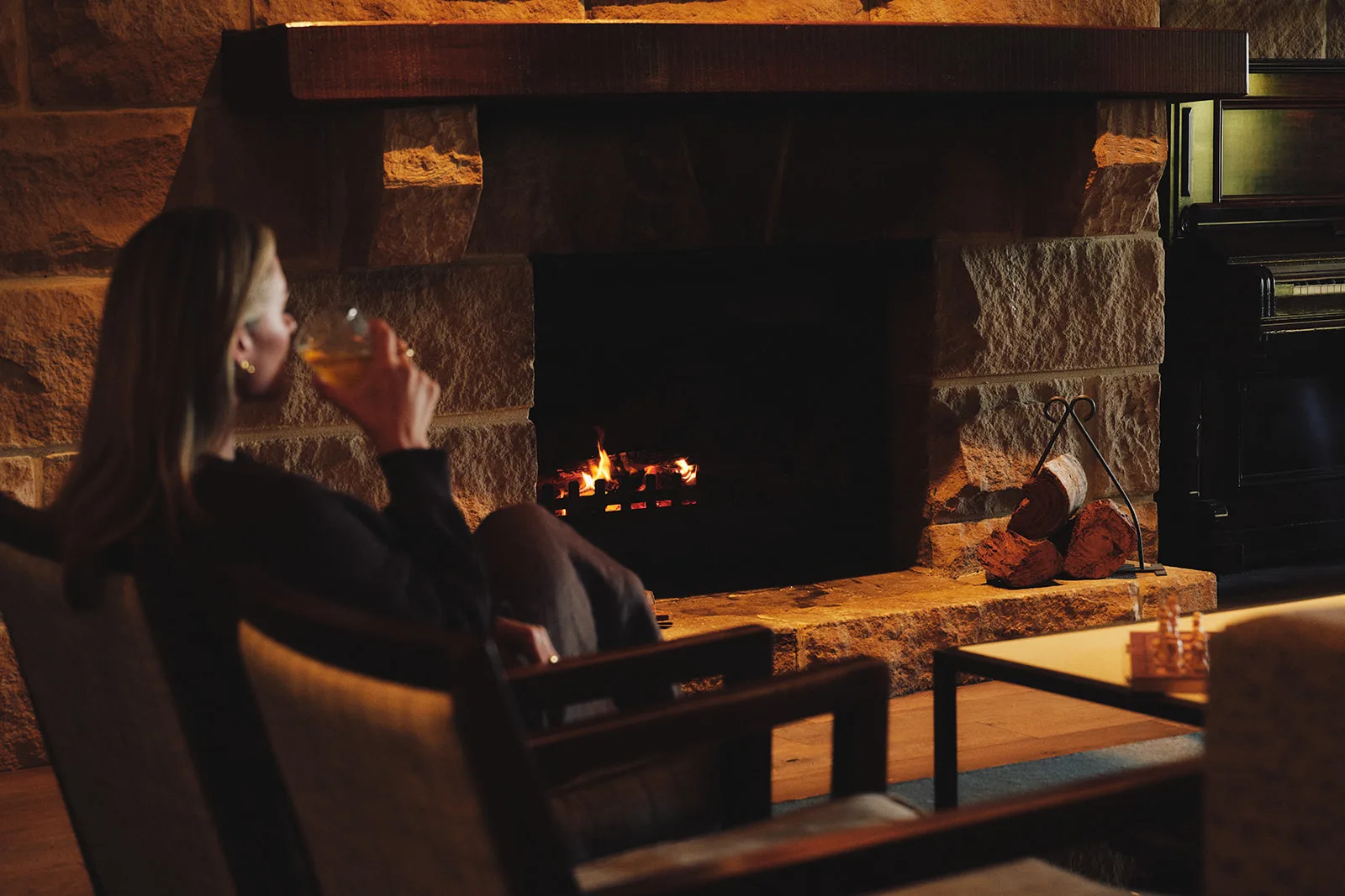 A woman sits by the fireplace with a mulled wine in her hand. 