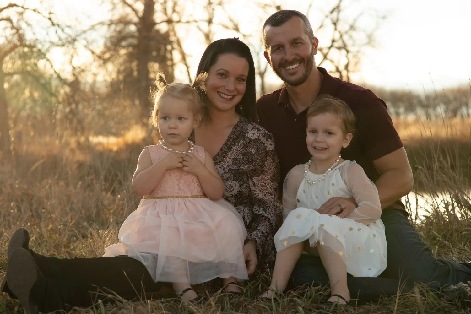 a potratit of a family in a sunny field, a mother father and two young girls smiling at the camera