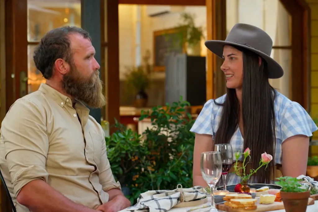 A man and a woman sitting across from each other at an outdoor dining table, appearing to be on a date. The man on the left has a full, thick beard and is wearing a beige button-down shirt, looking toward the woman. The woman on the right is smiling, wearing a grey wide-brimmed hat and a blue-and-white gingham dress. The table between them is set with wine glasses, a small vase with pink roses, and a platter of bread and cheese. The background shows a warmly lit indoor space through a doorway and lush green plants.