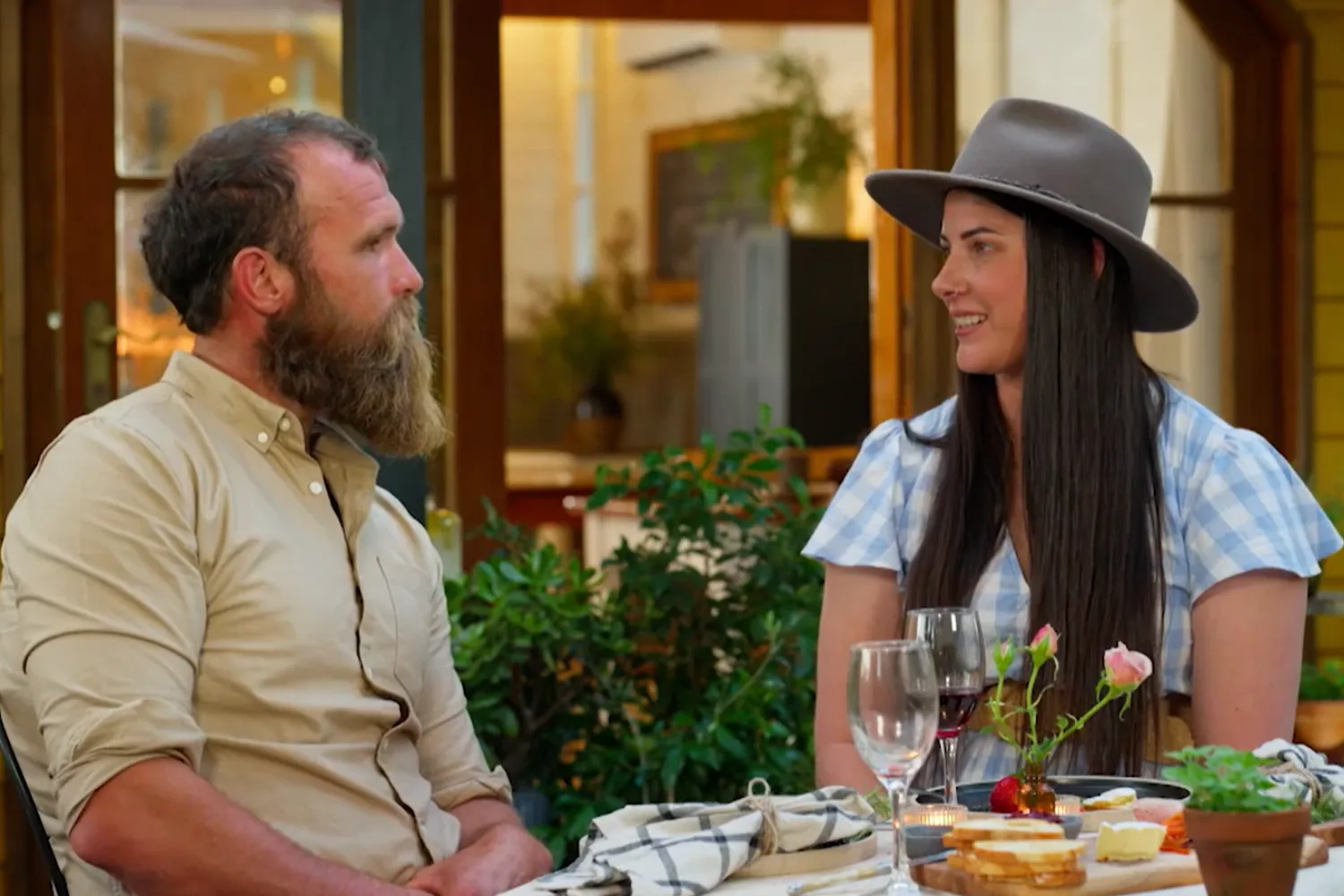 A man and a woman sitting across from each other at an outdoor dining table, appearing to be on a date. The man on the left has a full, thick beard and is wearing a beige button-down shirt, looking toward the woman. The woman on the right is smiling, wearing a grey wide-brimmed hat and a blue-and-white gingham dress. The table between them is set with wine glasses, a small vase with pink roses, and a platter of bread and cheese. The background shows a warmly lit indoor space through a doorway and lush green plants.