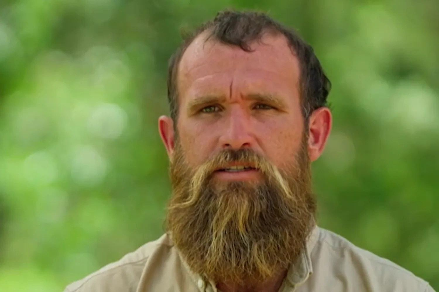 A close-up portrait of a man with a serious expression, looking directly forward. He has a full, prominent brown beard and mustache and is wearing a light beige button-down shirt. The background is a soft, completely blurred green foliage scene.