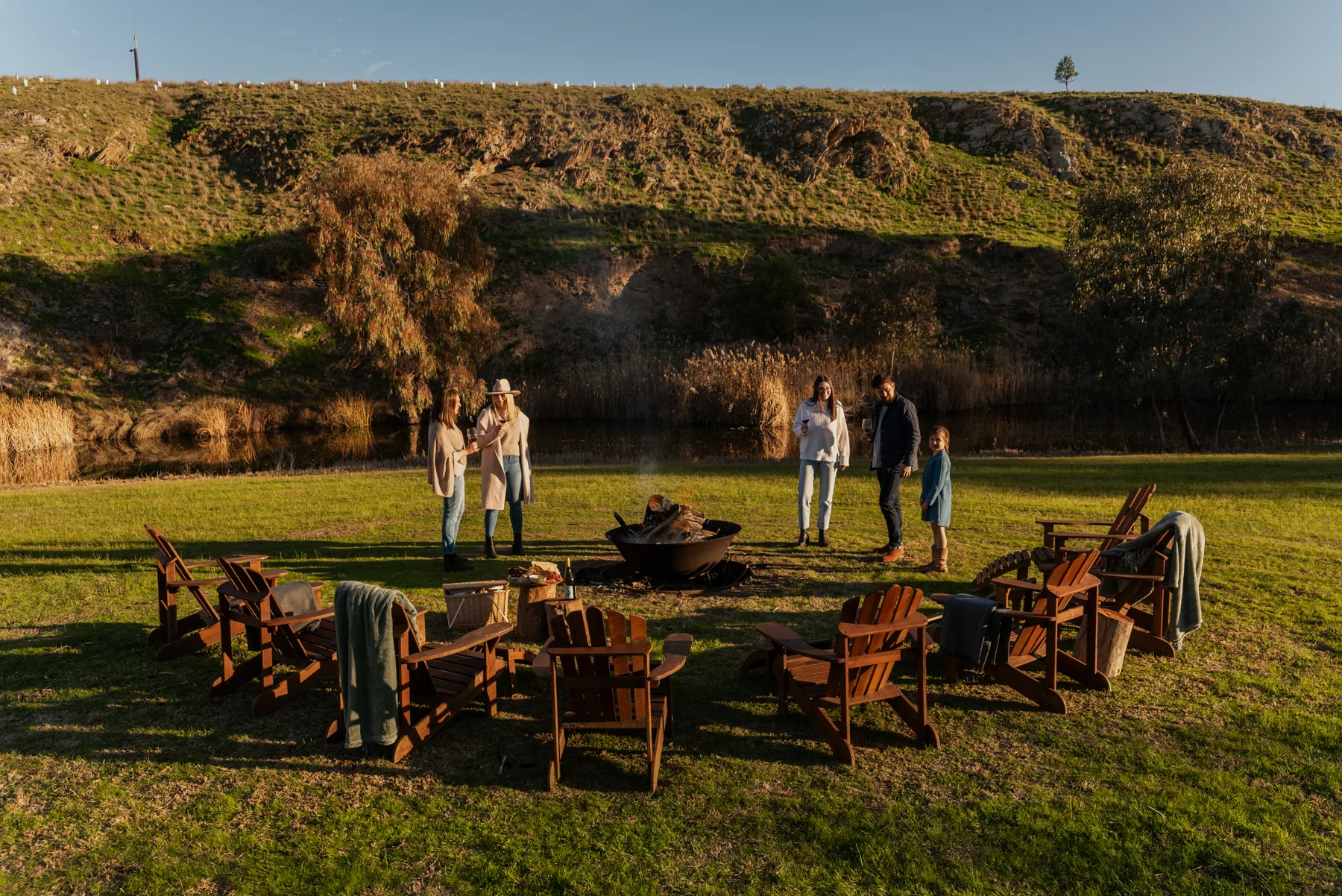 People mill about a firepit against rolling green hills. 