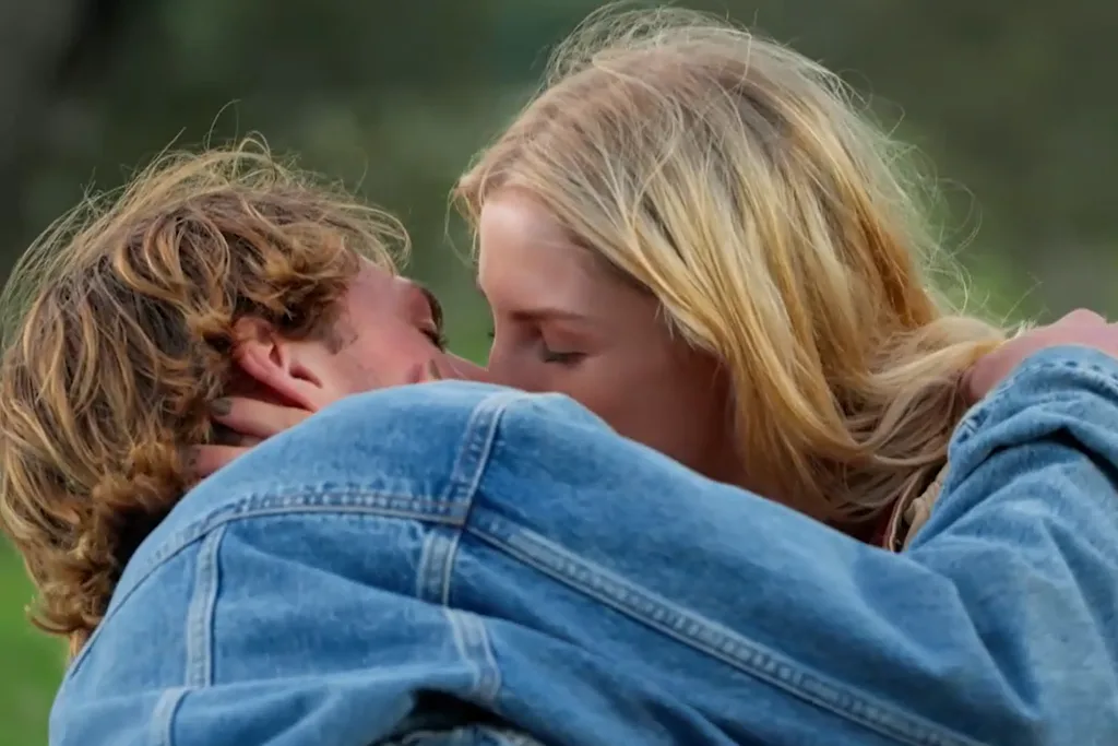 A sandy haired man in a denim jacket kisses a blonde woman.