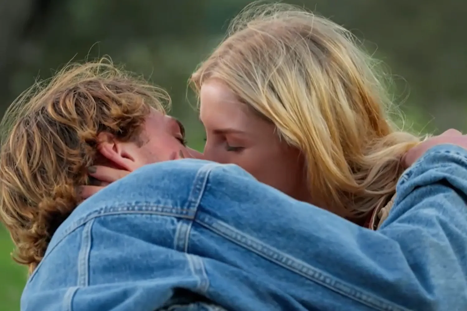 A sandy haired man in a denim jacket kisses a blonde woman. 