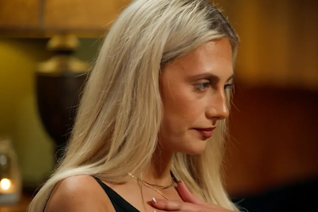 A blonde woman wearing a black tank top looks sad and shocked as she stands with her hand on her chest.