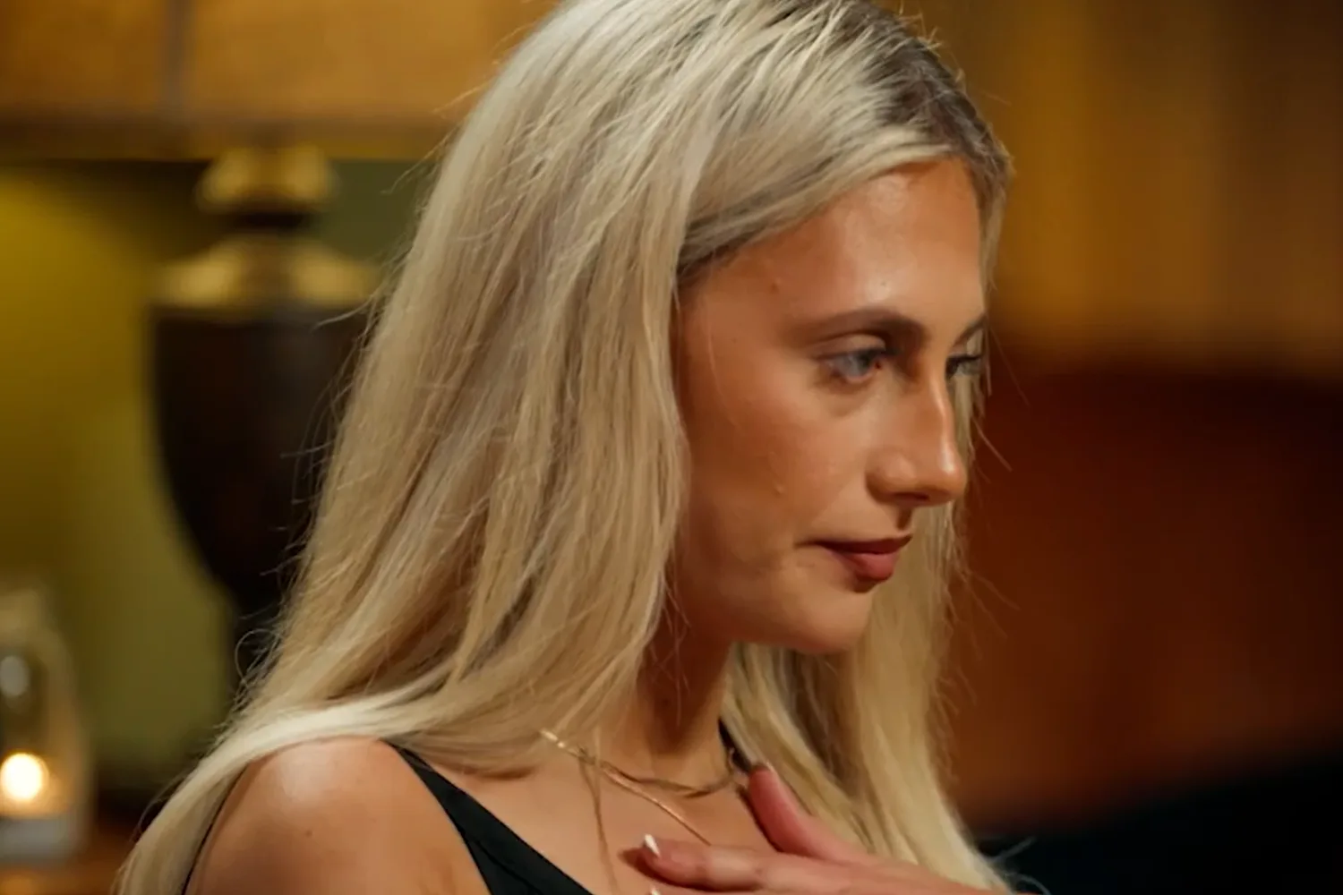 A blonde woman wearing a black tank top looks sad and shocked as she stands  with her hand on her chest. 