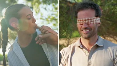 A split-screen image showing a playful outdoor blindfolded tasting game. On the left side, a woman with her hair in a long braid leans forward with a slight lens flare behind her, gently guiding another person's hand or tasting something from a finger. On the right side, a smiling man with a short beard wears a brown-and-white checkered blindfold, waiting expectantly. Both are outdoors with soft-focus green foliage in the background.
