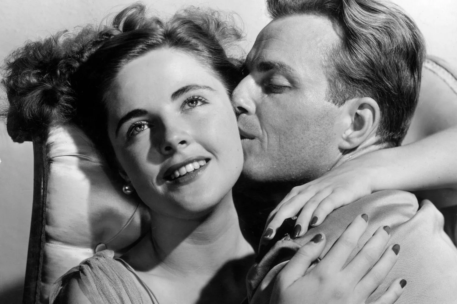 Headshot of a woman smiling and embracing a man as he kisses her on the cheek.