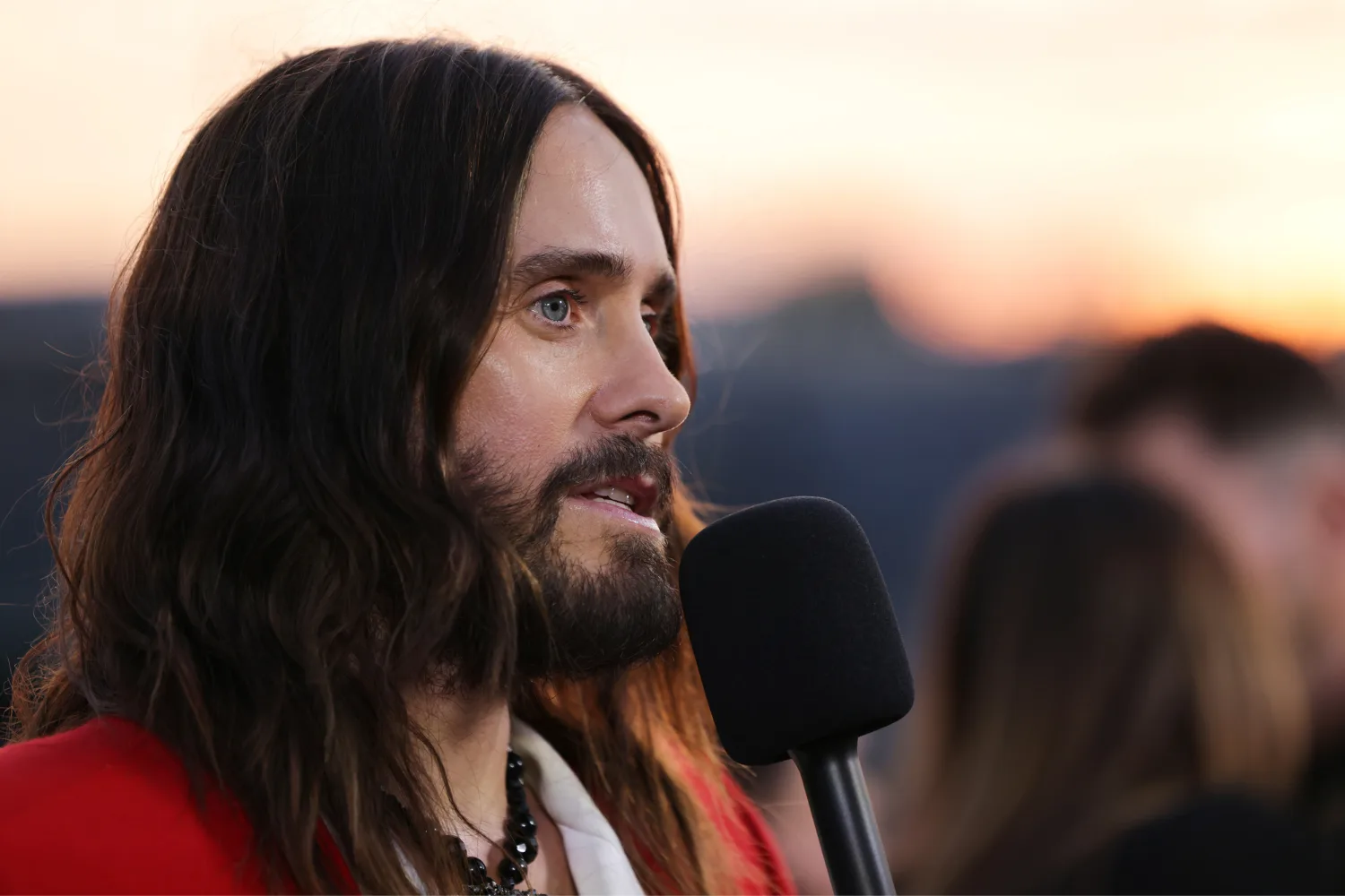 Jared Leto speaks into a microphone during a red carpet interview at an event. He has shoulder-length dark hair and a beard, and is wearing a red jacket over a white shirt with layered necklaces. The background is softly blurred with other attendees and a sunset sky.