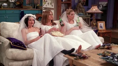 Three women in wedding dresses and veils sit on a couch, eating popcorn and drinking beer while watching TV.
