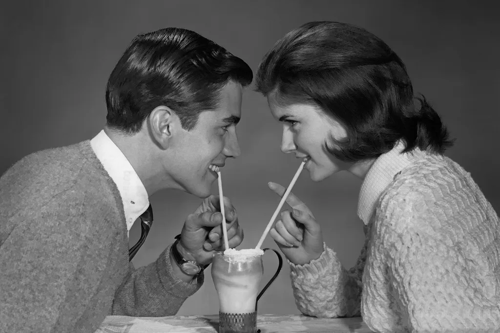 Smartly dressed young couple facing each other share a milkshake.