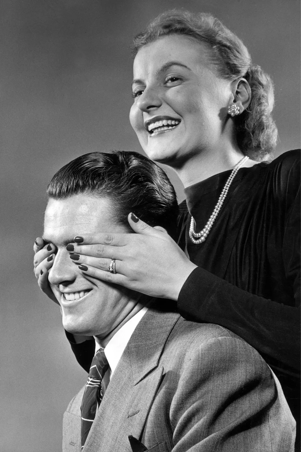  A smiling woman, wearing pearl earrings and a pearl necklace, stands and covers the eyes of a seated man.