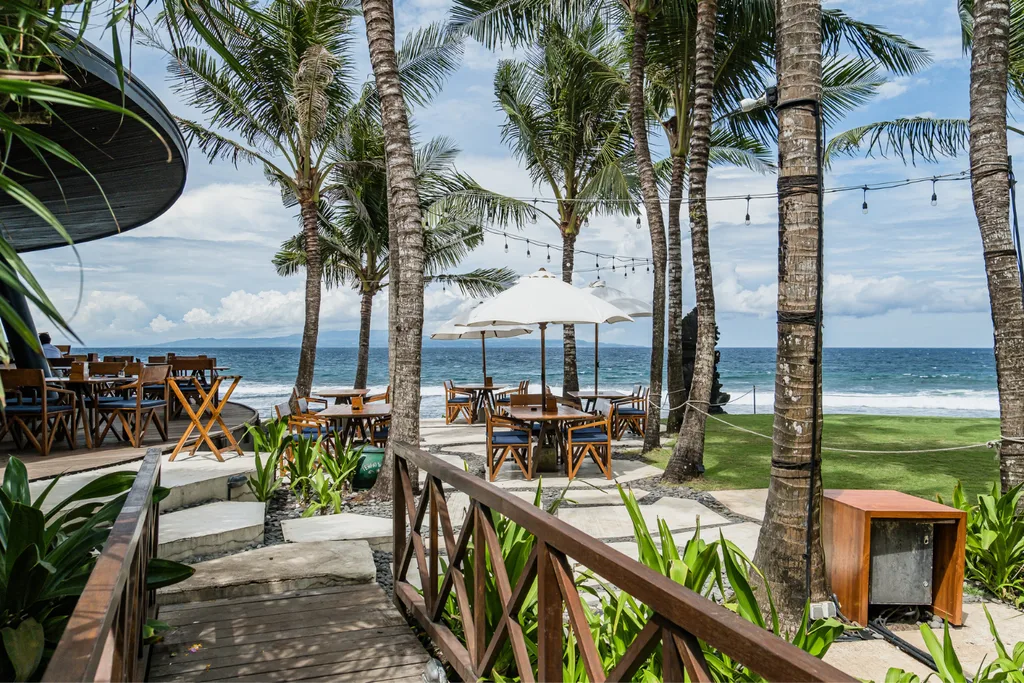 An outdoor beachfront restaurant with wooden tables, director-style chairs, and white umbrellas set among tall palm trees overlooking a blue ocean under a party-cloudy sky. A small wooden bridge with a crisscross railing leads into the dining area.