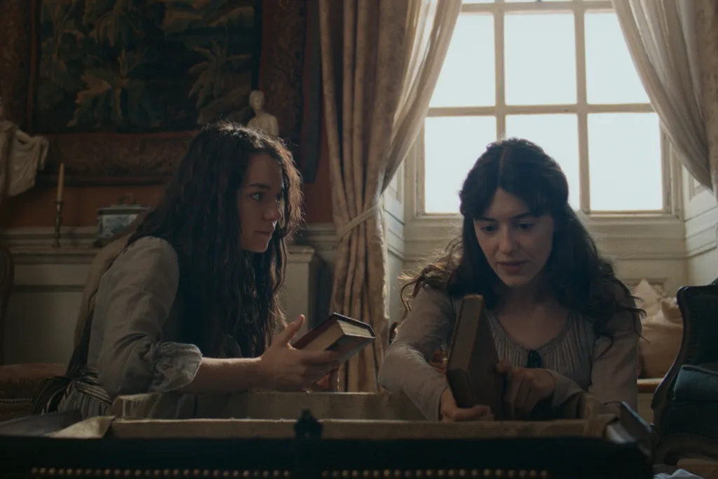 A scene from a period drama featuring two young women in an elegantly furnished room, leaning over an open wooden chest while examining books. The woman on the left has long, wavy brown hair and wears a cream blouse with rolled-up sleeves and a striped skirt. She holds an open book and looks towards her companion. The woman on the right has dark brown hair styled half-up and wears a pale grey dress with delicate detailing around the neckline. She holds a brown hardcover book and points towards something inside the chest. Behind them, a large multi-paned window framed by heavy cream curtains lets in soft natural light. Antique furnishings, decorative artwork and warm, muted tones create an intimate, historical atmosphere.