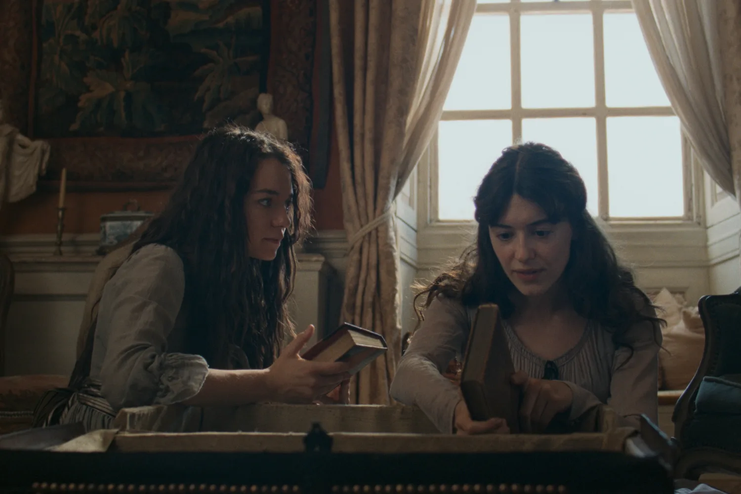 A scene from a period drama featuring two young women in an elegantly furnished room, leaning over an open wooden chest while examining books. The woman on the left has long, wavy brown hair and wears a cream blouse with rolled-up sleeves and a striped skirt. She holds an open book and looks towards her companion. The woman on the right has dark brown hair styled half-up and wears a pale grey dress with delicate detailing around the neckline. She holds a brown hardcover book and points towards something inside the chest. Behind them, a large multi-paned window framed by heavy cream curtains lets in soft natural light. Antique furnishings, decorative artwork and warm, muted tones create an intimate, historical atmosphere.