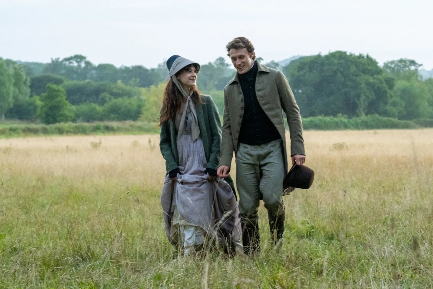 A romantic scene from a period drama showing a young couple walking together through a sprawling meadow of tall, golden grass. The woman wears a pale lavender-grey dress, a dark green coat and a blue-grey bonnet tied beneath her chin. She gathers the fabric of her long skirt in her hands while smiling up at the man beside her. He wears a sage-green Regency-era suit with a black waistcoat, high-collared shirt and tall black boots, carrying a black top hat in one hand. The pair exchange affectionate smiles as they stroll through the countryside, surrounded by lush green trees beneath an overcast sky.