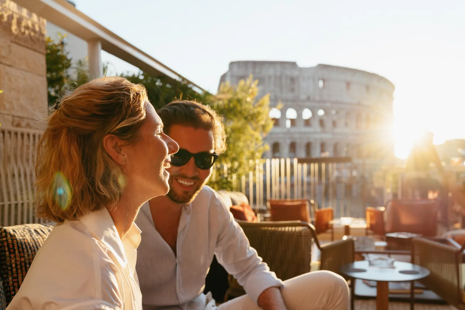 A smiling couple sitting on a sunny rooftop terrace in Rome, with the Colosseum visible in the background.