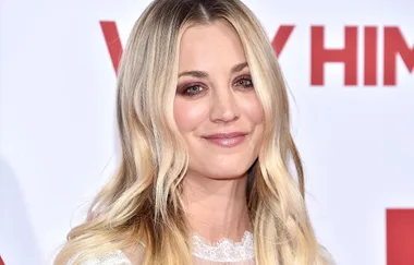 A person with long blonde hair smiles at a red carpet event, wearing a white lace outfit, against a blurred backdrop.