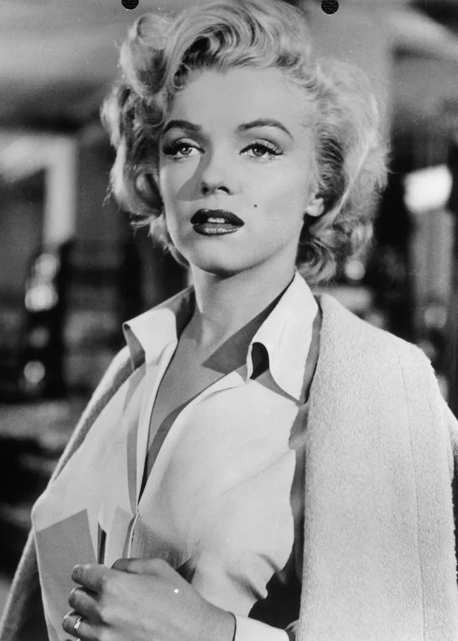 A woman with short blond hair and a white blouse gazes upward, with soft lighting highlighting her features.