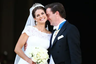 Wedding couple smiling, bride in lace gown and tiara holding bouquet, groom in formal suit with medals, embracing closely.
