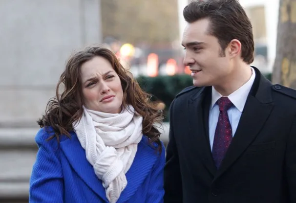 A woman in a blue coat and white scarf makes a funny face while walking with a man in a suit and red tie outdoors.
