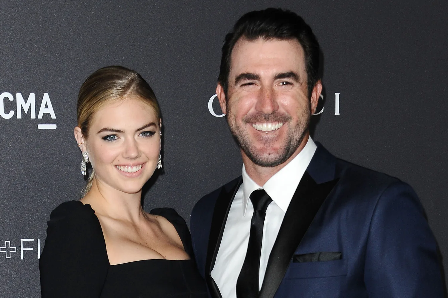A woman in a black dress and a man in a suit smile at a formal event with a branded backdrop.