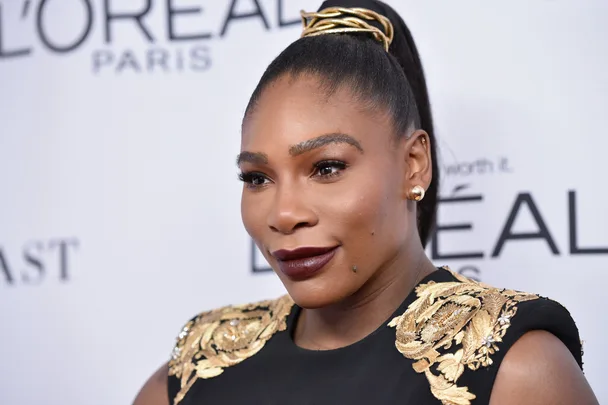 A woman in a black and gold embroidered dress at a L'Oreal Paris event, with a high ponytail and dark lipstick.