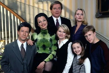 A group photo of seven people posing indoors, three standing and four seated, in front of a staircase with striped wallpaper.