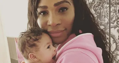 A woman in a pink hoodie holds a baby close, both looking at the camera.
