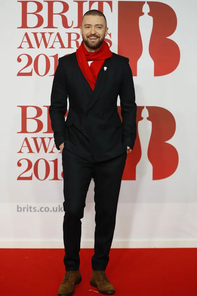 Man in a black suit and red scarf smiling on the red carpet at the Brit Awards 2018.