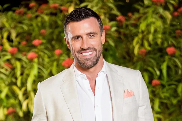 A man in a light suit smiles in front of a lush green background with red flowers.